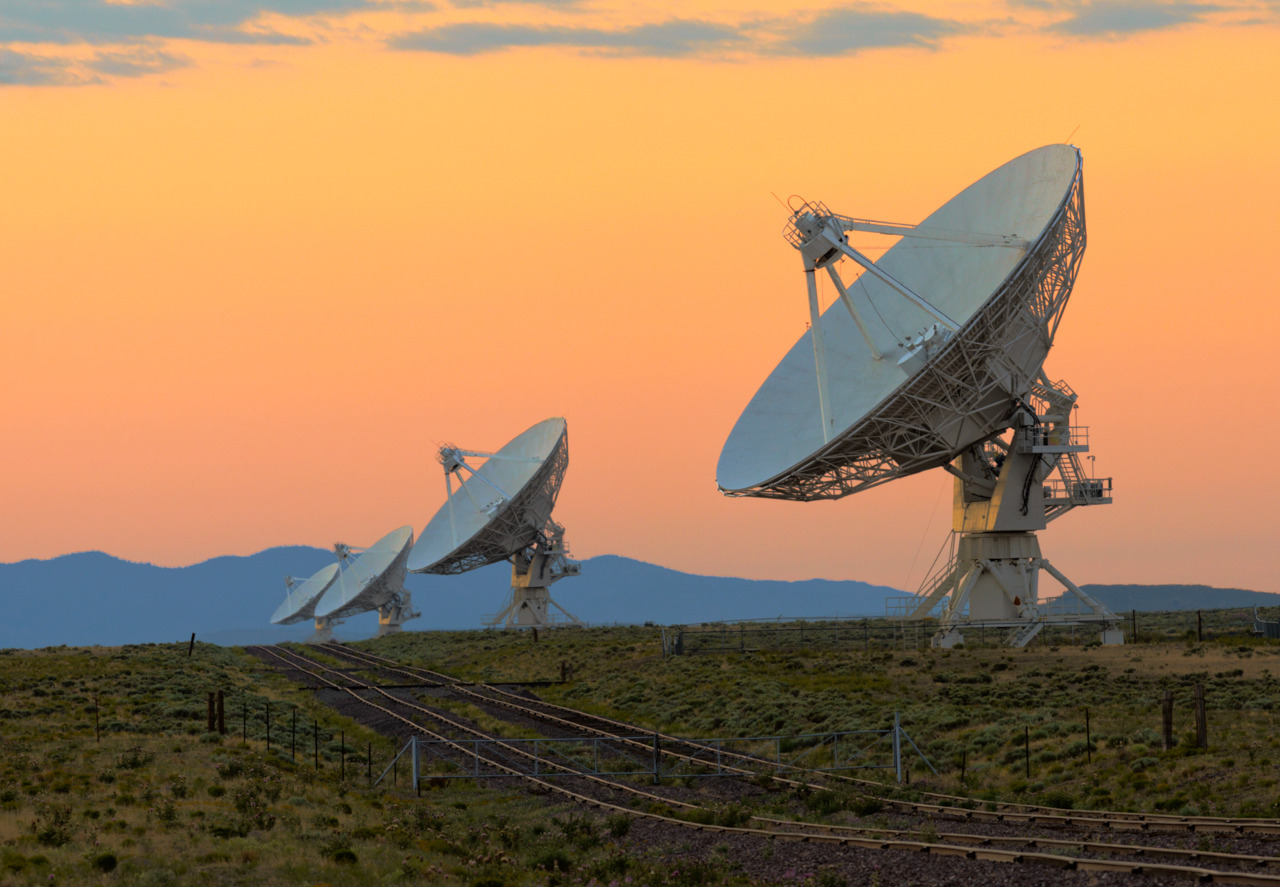 Row of large satelite dishesin a filed at sunset with rail tracks.